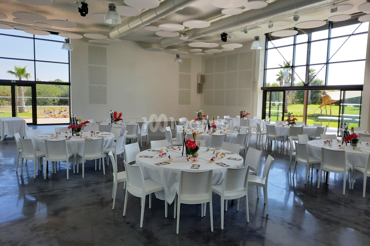 Salle lumineuse avec tables rondes dressées, chaises blanches et décoration florale, donnant sur un jardin extérieur.