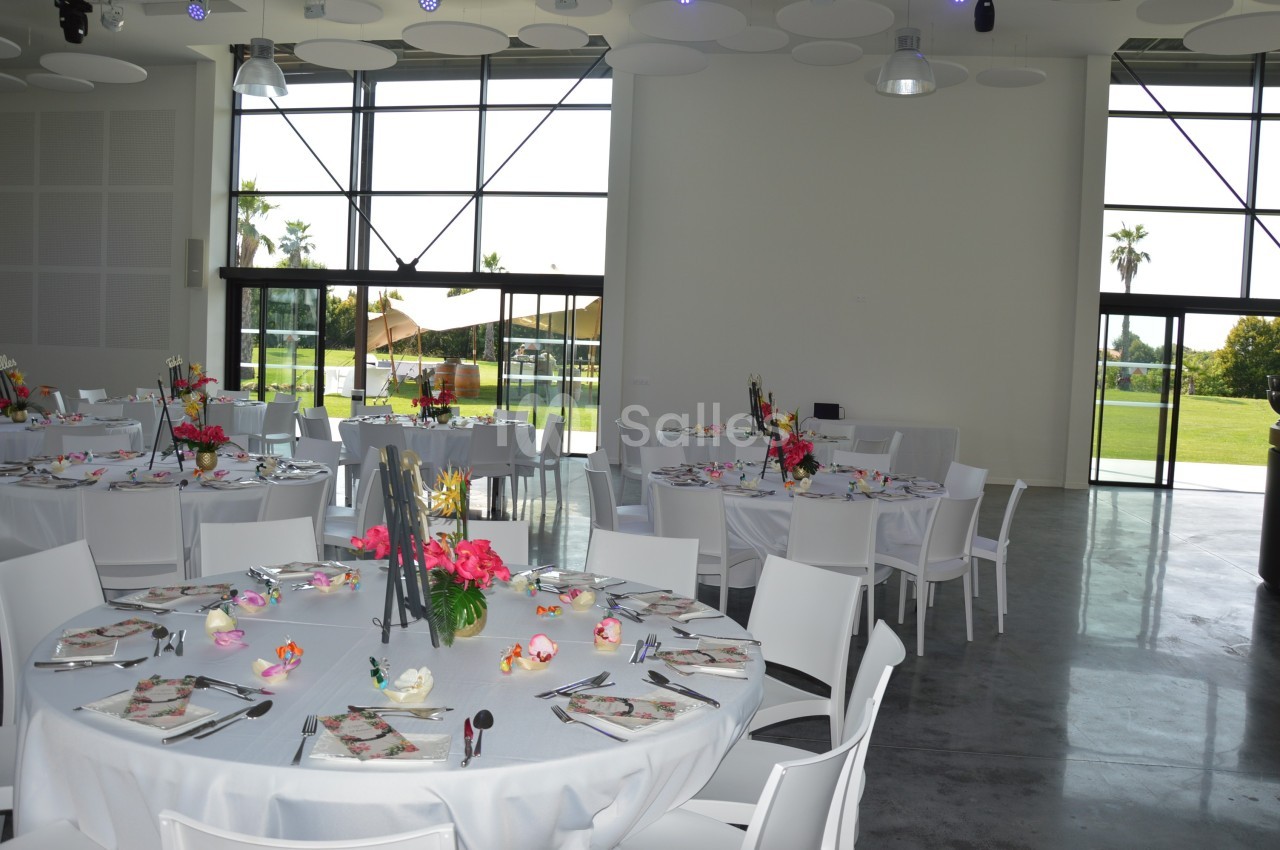 Salle lumineuse avec tables rondes dressées pour un événement, décorées de fleurs colorées, vue sur un jardin extérieur.