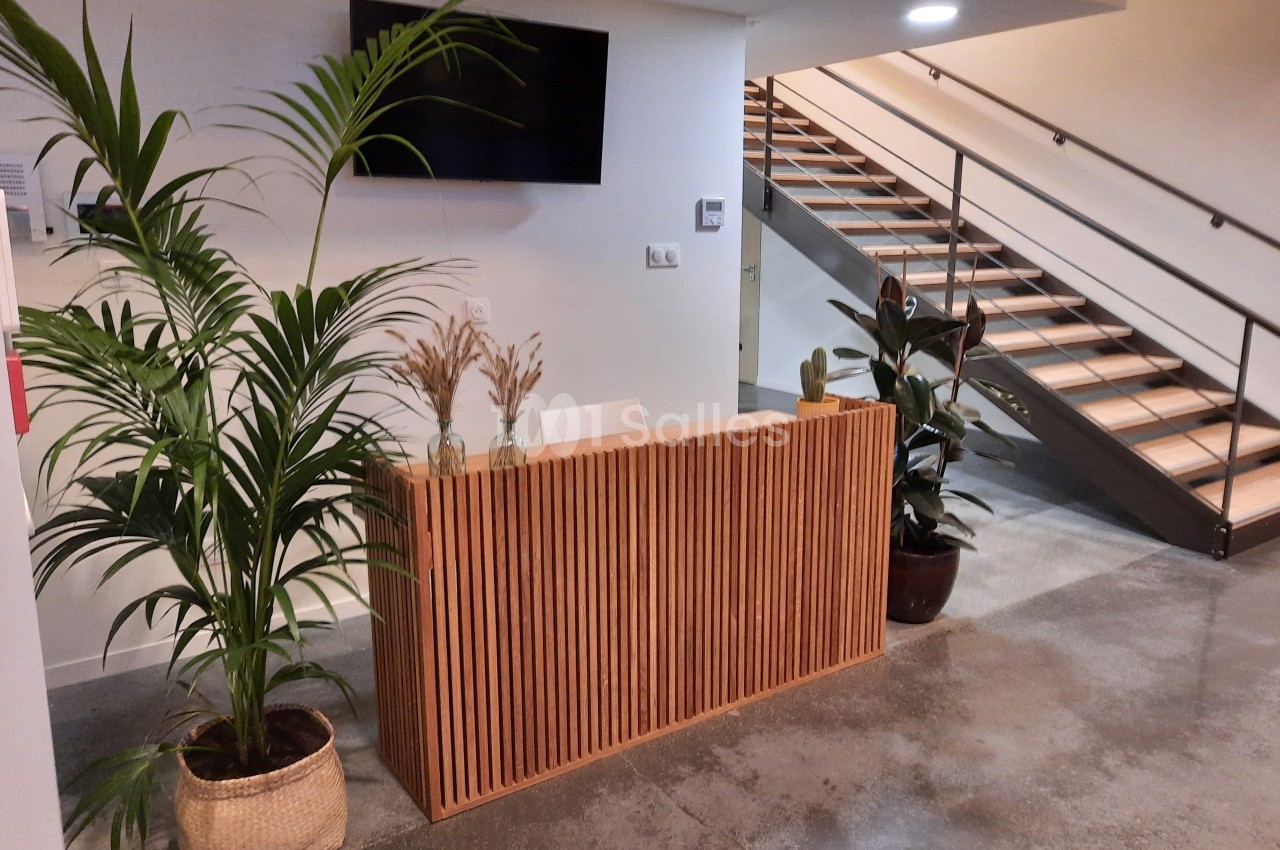 Accueil avec un comptoir en bois, plantes décoratives, escalier en arrière-plan et écran mural.