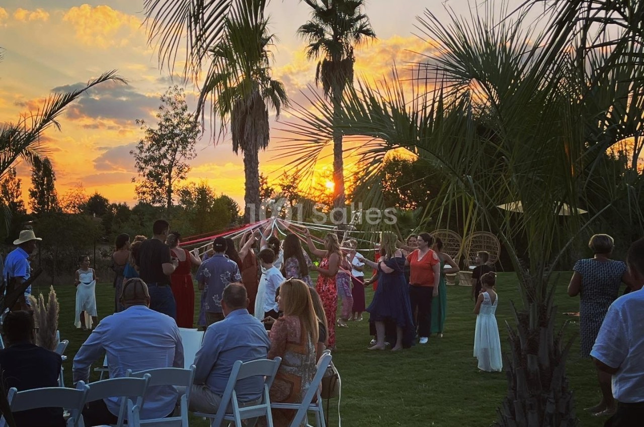 Groupe de personnes célébrant en plein air au coucher du soleil, entouré de palmiers et de chaises disposées sur une pelouse.