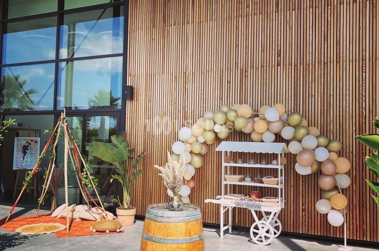 Décoration extérieure avec arche de ballons, tipi, tonneau en bois et étagère sur fond de façade en bois et verre.