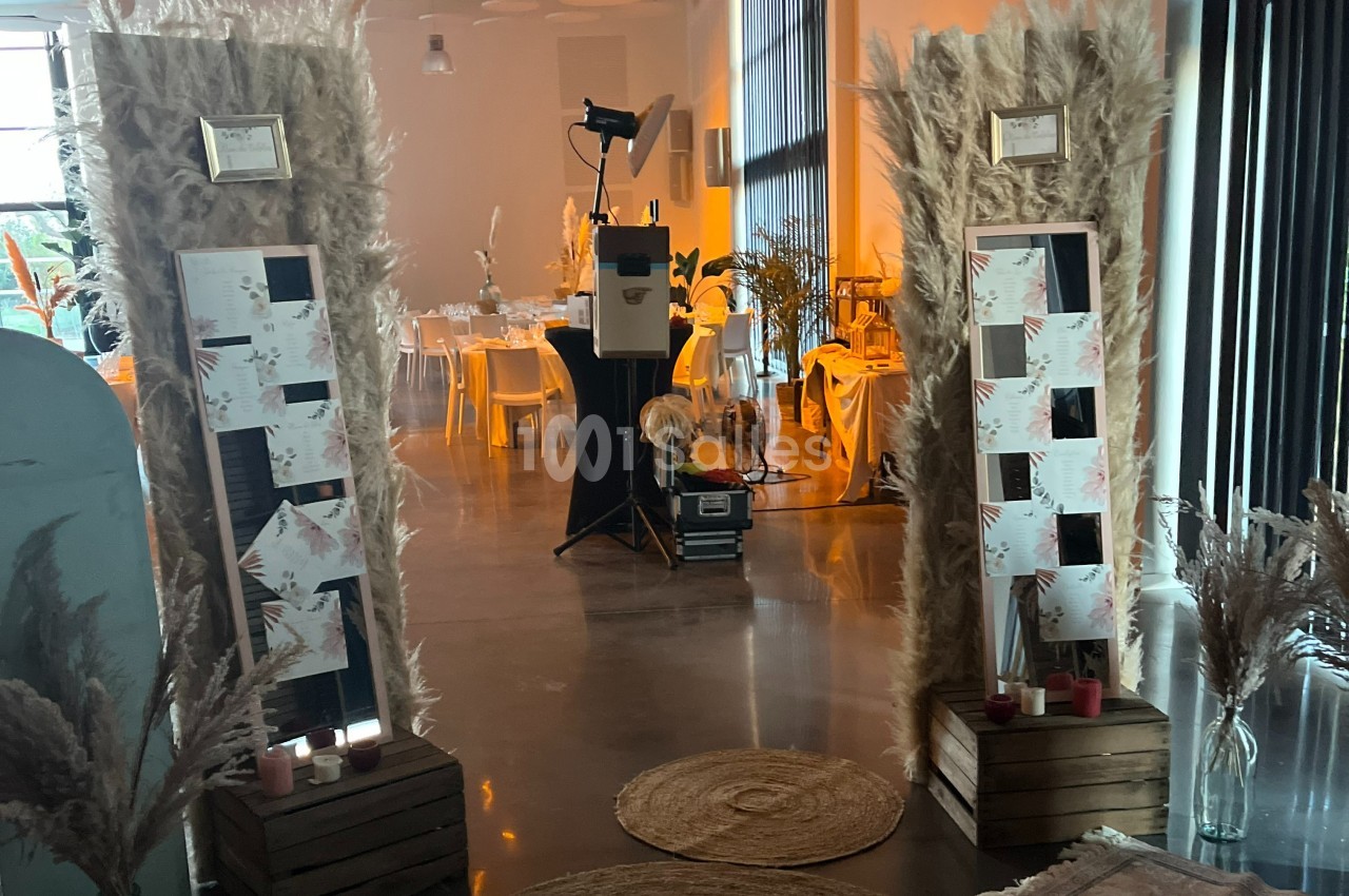 Deux panneaux décorés de pampas et cartes, encadrant une entrée dans une salle lumineuse avec tables dressées.