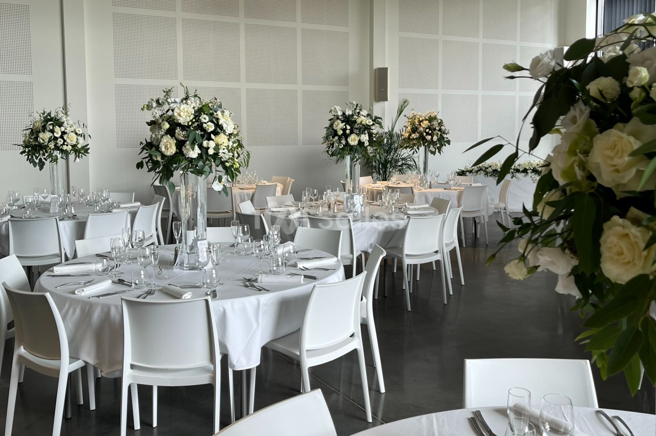 Salle de réception élégante avec tables rondes dressées, nappes blanches et arrangements floraux blancs.