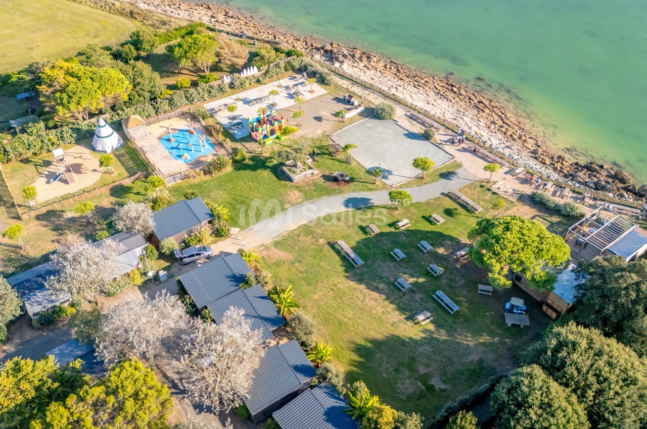 Vue aérienne d'un camping en bord de mer avec espaces verts, hébergements, aire de jeux et accès à une plage rocheuse.