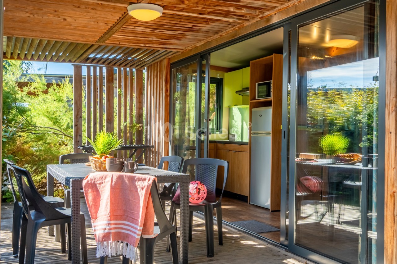 Terrasse en bois couverte avec table, chaises et vue sur une cuisine équipée à travers des portes vitrées.