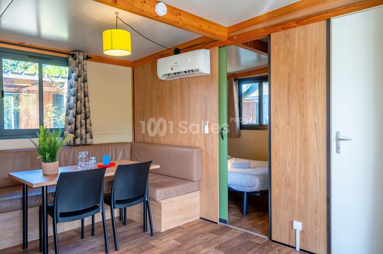 Intérieur d'un mobil-home avec coin repas, canapé, table, chaises, et une chambre visible en arrière-plan.