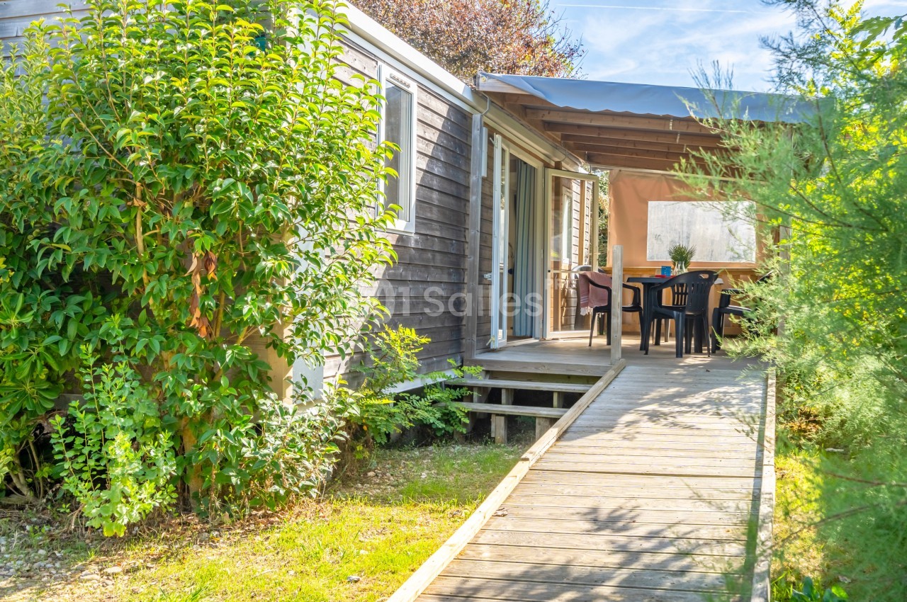 Mobil-home avec terrasse en bois couverte, entouré de végétation, sous un ciel dégagé.