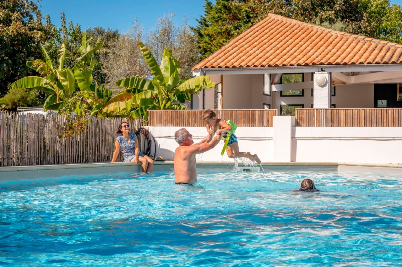 Un homme soulève un enfant au-dessus de l'eau dans une piscine extérieure, avec des personnes en arrière-plan.