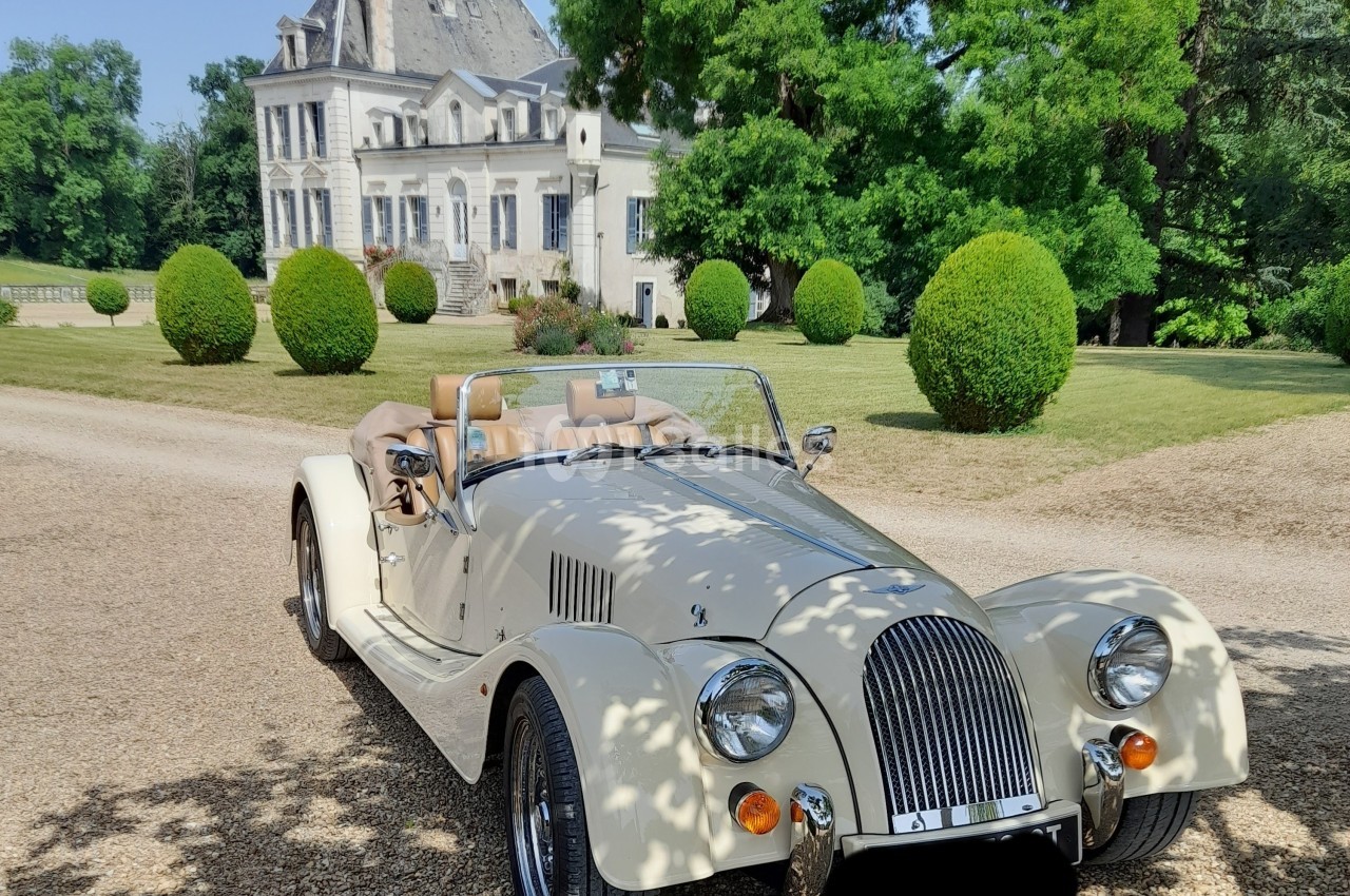 Voiture de collection décapotable beige stationnée sur une allée gravillonnée devant un manoir entouré de verdure.
