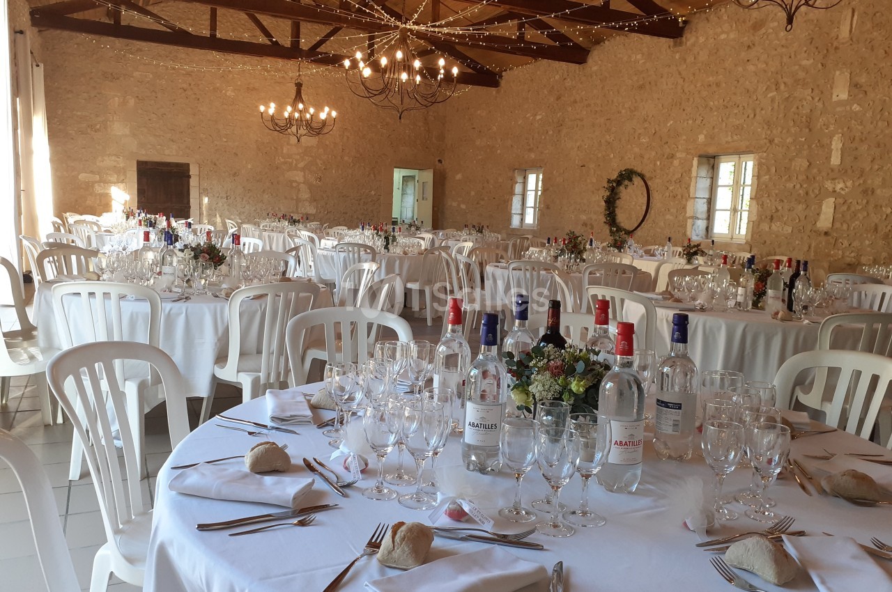 Salle de réception en pierre avec tables rondes dressées, nappes blanches, chaises en plastique et décorations lumineuses.