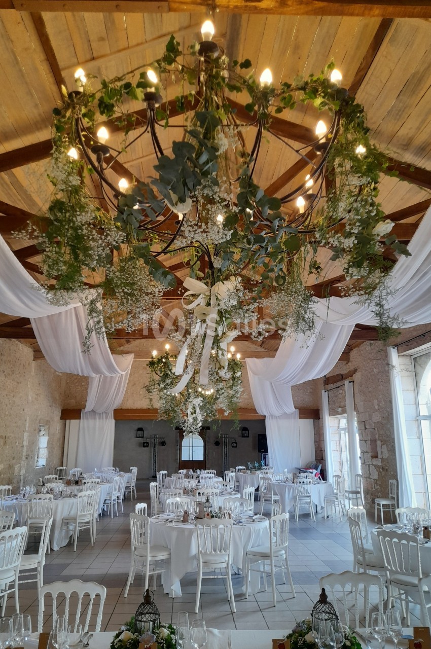 Salle de réception décorée avec des tables dressées, des chaises blanches et un plafond orné de guirlandes végétales.
