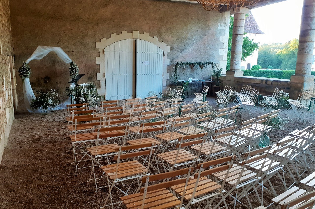 Chaises en bois disposées pour une cérémonie dans une cour couverte avec arche florale et vue sur un jardin.