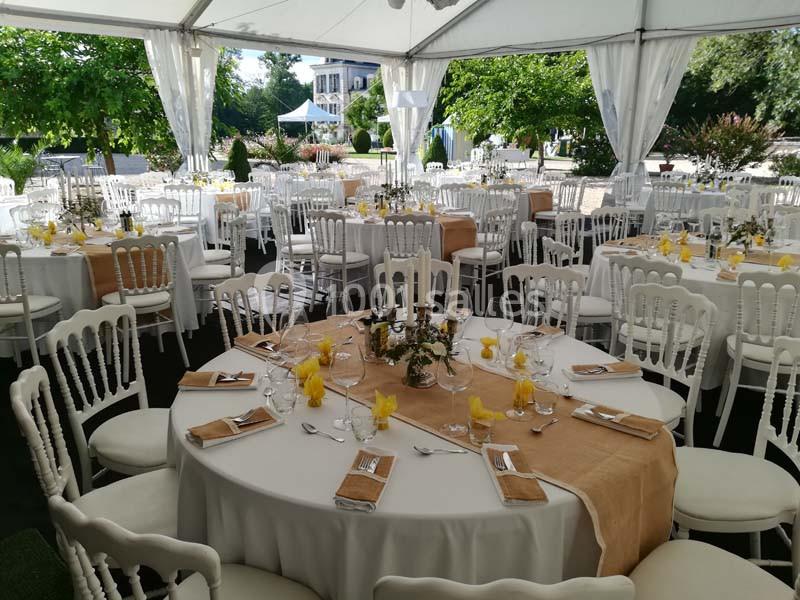 Tables rondes décorées pour un événement, disposées sous une tente avec nappes blanches et chemins de table beige.