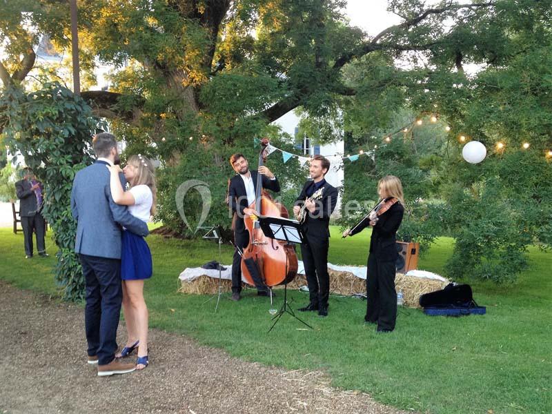 Un groupe de musiciens joue en plein air près d'un couple dansant sur une pelouse, entourés d'arbres et de guirlandes…