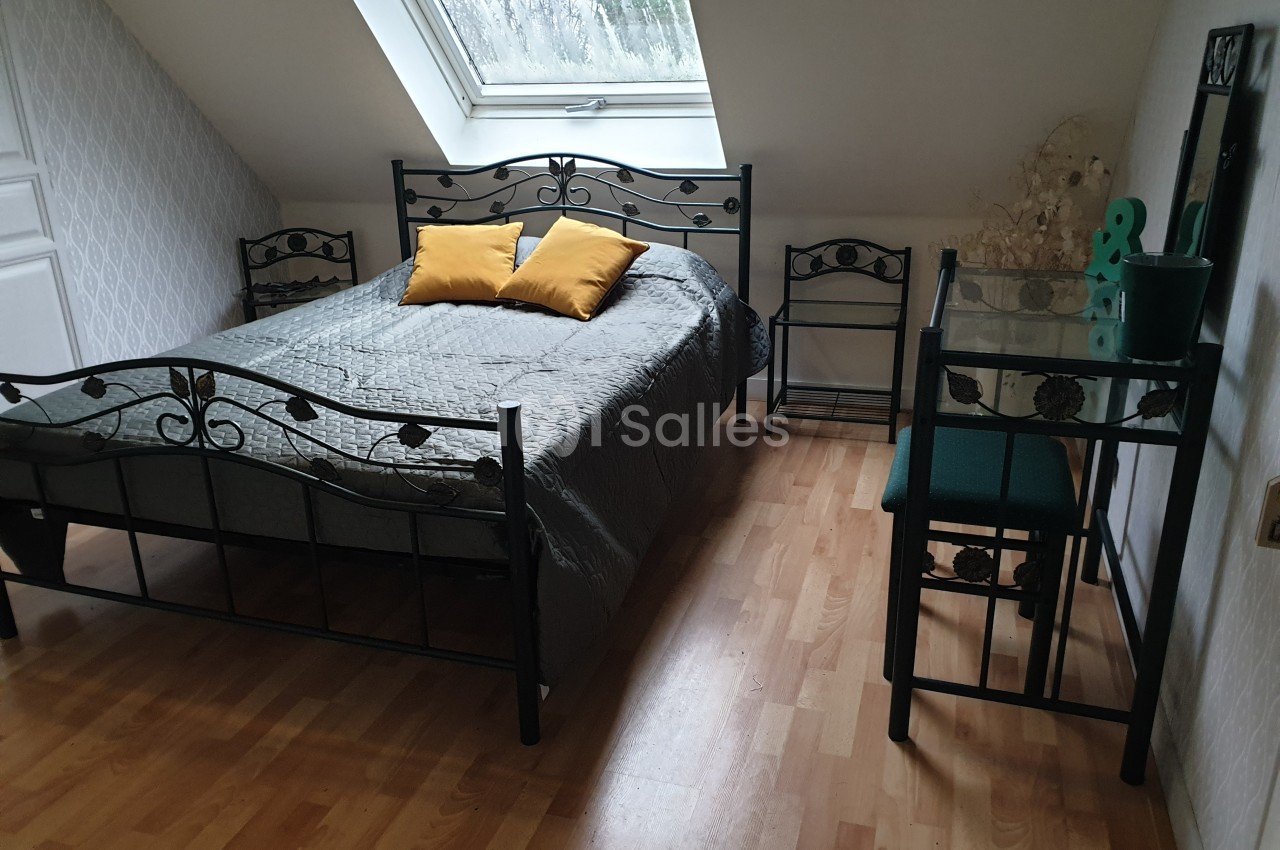Chambre mansardée avec lit double en métal noir, coussins jaunes, bureau en verre et parquet clair.