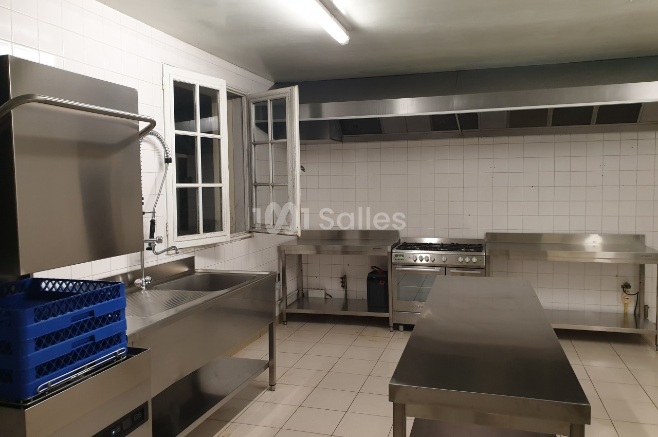 Cuisine professionnelle avec équipements en inox, plan de travail, évier, four, hotte et fenêtre ouverte.