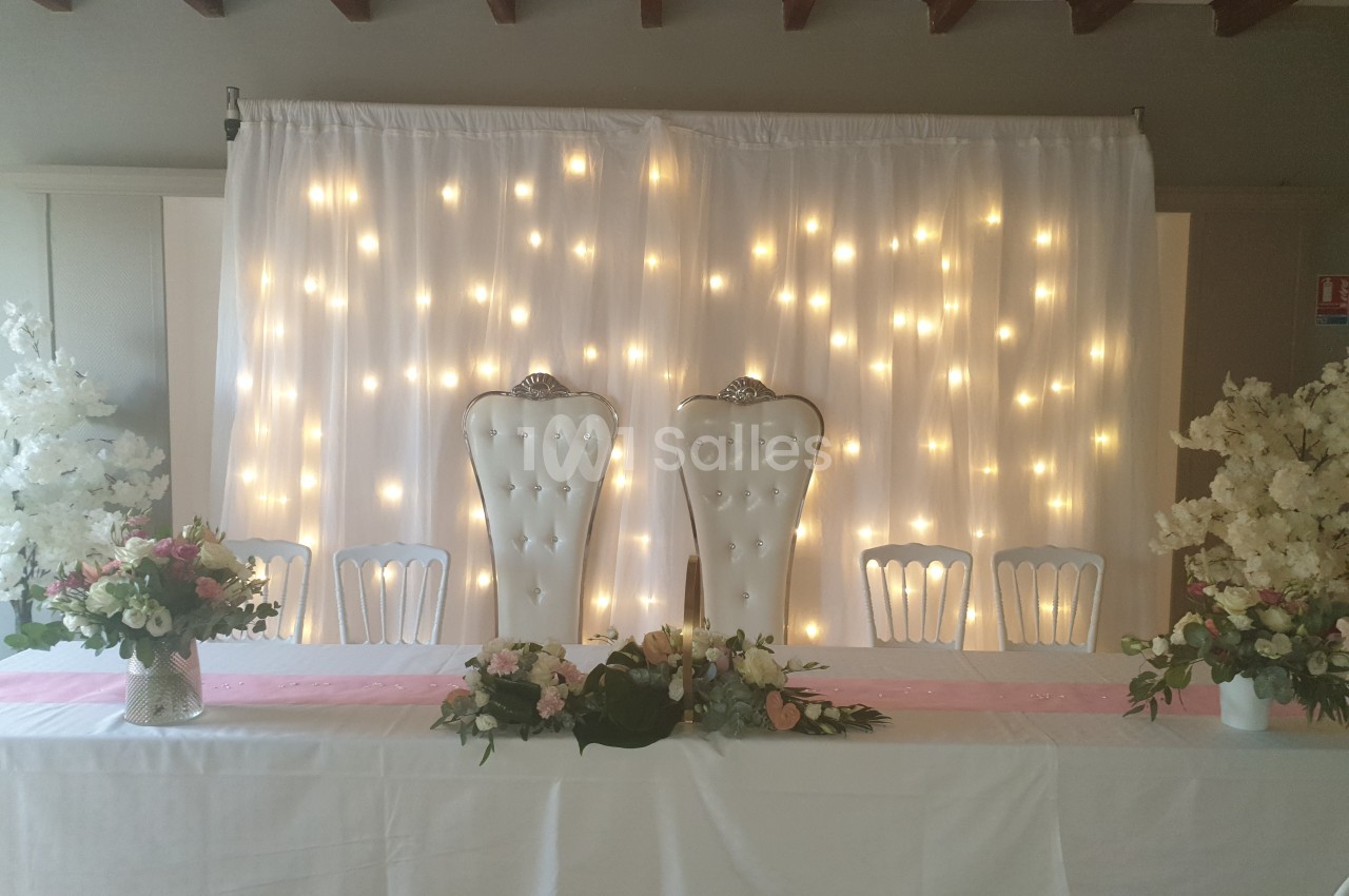 Salle décorée pour un mariage avec deux fauteuils blancs capitonnés, guirlandes lumineuses et arrangements floraux.