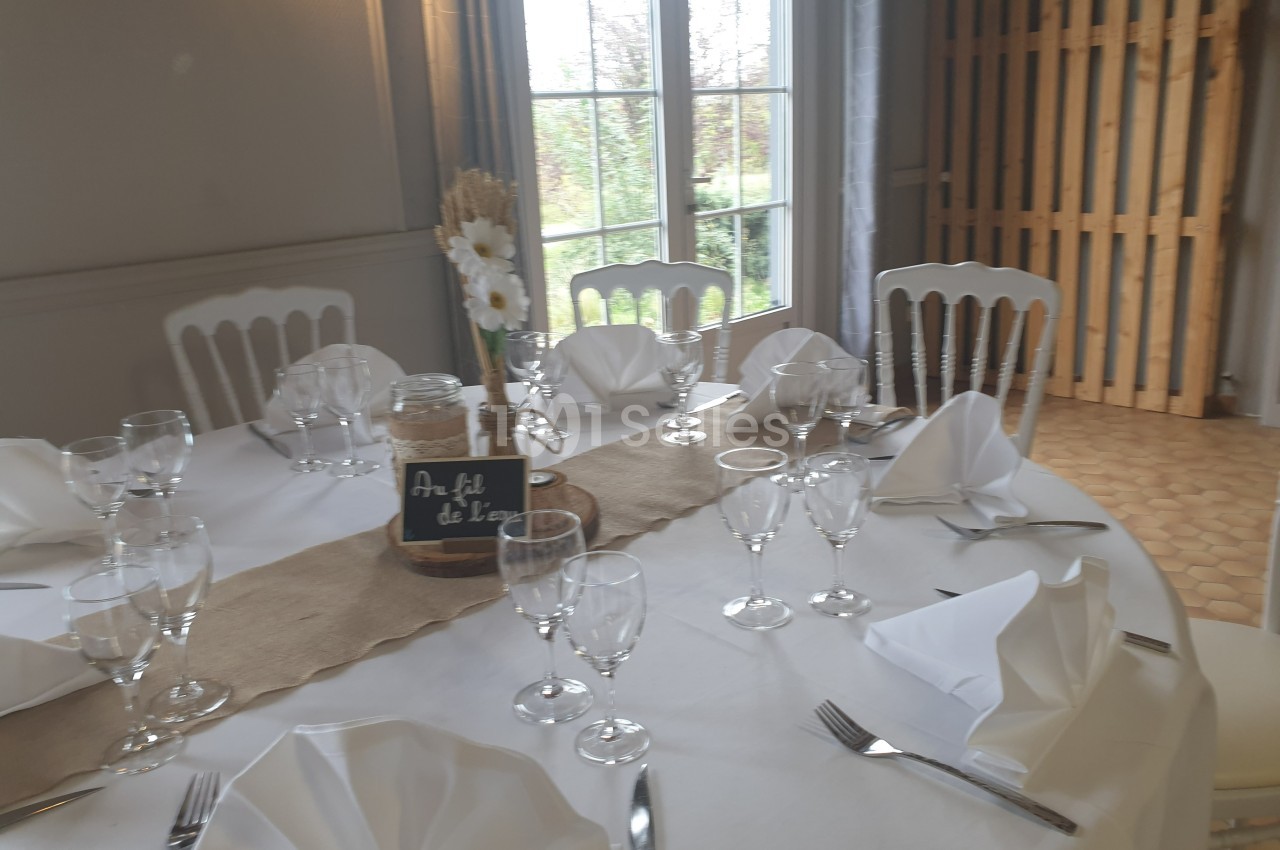 Table ronde dressée avec nappes blanches, serviettes pliées, verres et couverts, décorée de fleurs et d'une ardoise.