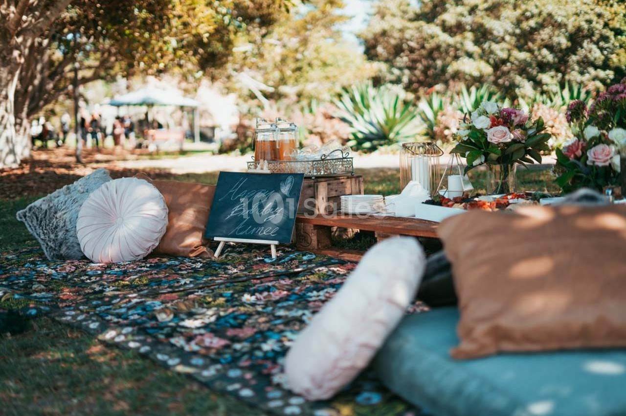 Pique-nique en plein air avec coussins, tapis coloré, table basse garnie de boissons, fleurs et ardoise décorative.