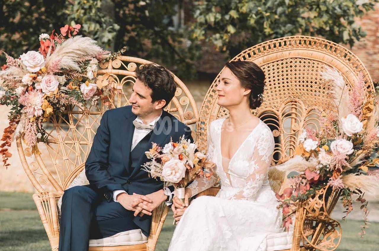 Un couple en tenue de mariage est assis sur des fauteuils en rotin décorés de fleurs, en extérieur.