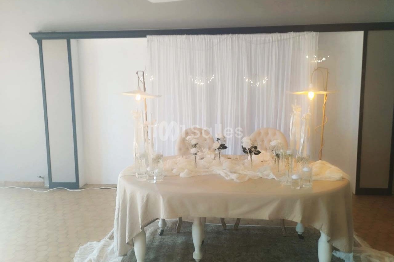 Table décorée pour un événement avec nappes beiges, chaises capitonnées, fleurs blanches et éclairage doux.