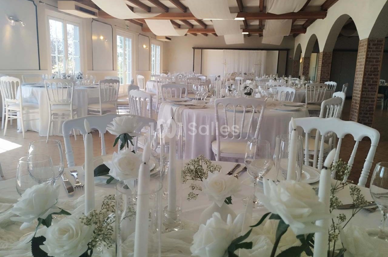 Salle de réception décorée pour un événement, avec des tables rondes dressées et ornées de fleurs blanches.