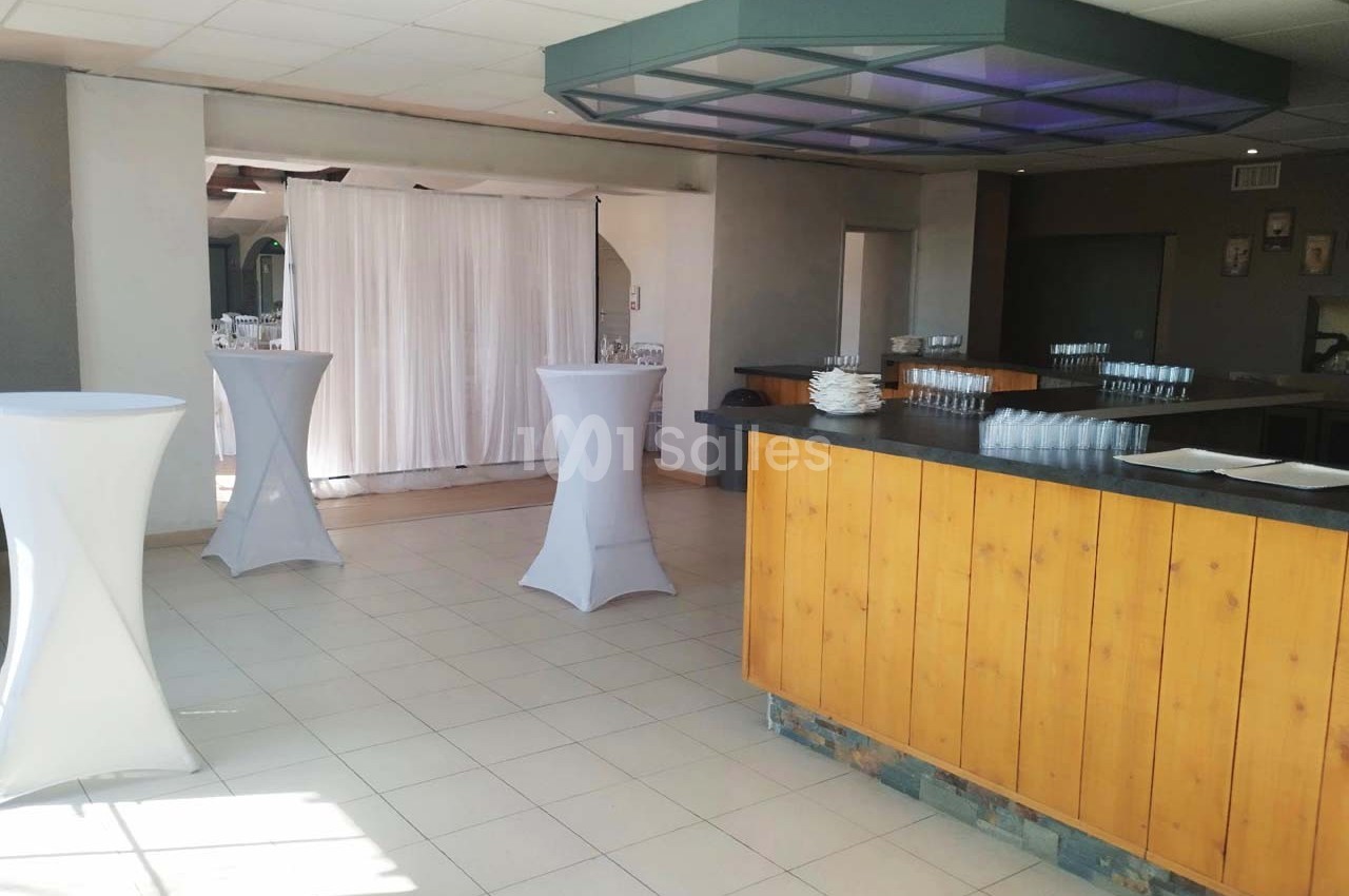 Salle lumineuse avec un bar en bois, des tables hautes drapées de blanc et un rideau blanc en arrière-plan.