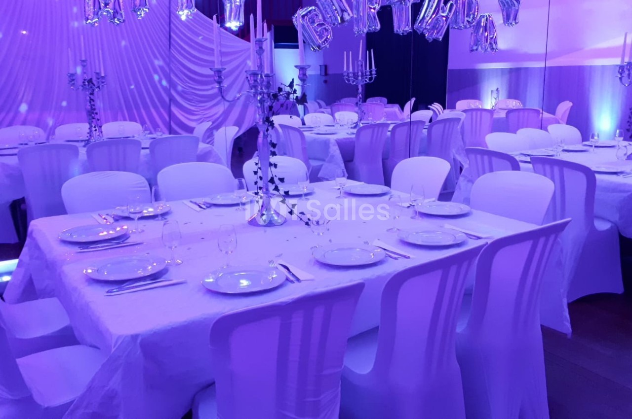 Salle décorée pour un événement, avec tables dressées, nappes blanches, chandeliers et éclairage violet.