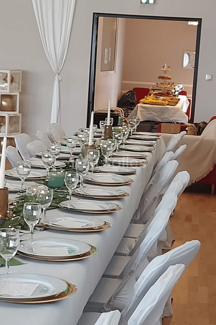 Table dressée avec nappes blanches, assiettes, verres et bougies, dans une salle avec buffet visible en arrière-plan.