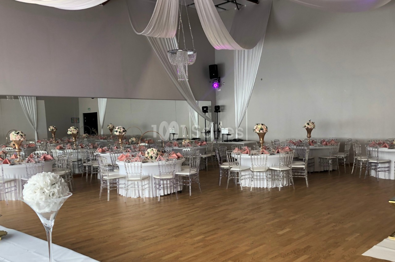 Salle de réception décorée avec des tables rondes, nappes blanches, chaises transparentes et arrangements floraux.