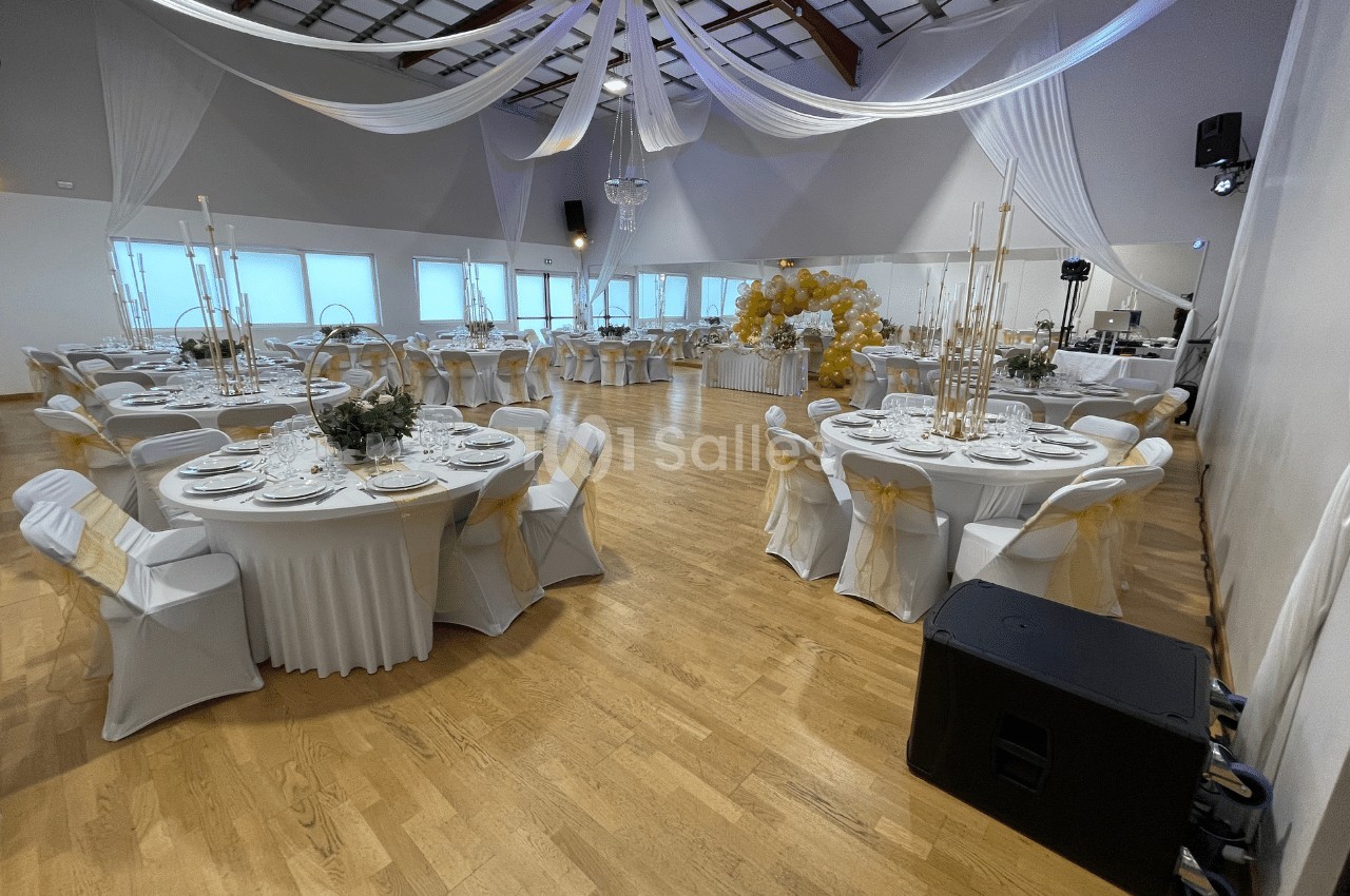 Salle de réception décorée avec des tables rondes dressées, nappes blanches et chaises ornées de rubans dorés.