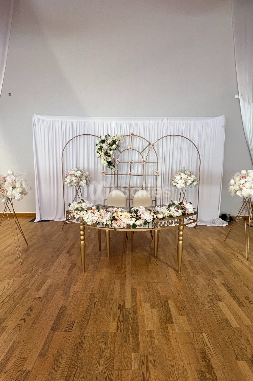 Décoration de mariage avec table ornée de fleurs blanches et roses, chaises blanches et arche métallique dorée.
