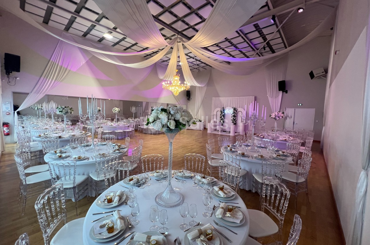 Salle de réception décorée avec des tables rondes dressées, des chaises transparentes et des drapés suspendus au plafond.