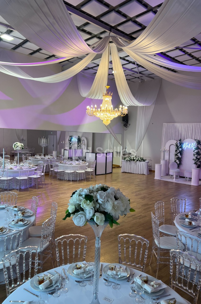 Salle de réception élégante avec tables décorées, chaises transparentes, lustre central et drapés suspendus au plafond.