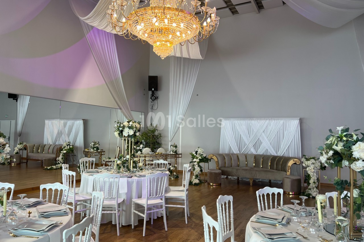Salle de réception élégante avec tables dressées, chaises blanches, décor floral et grand lustre central.