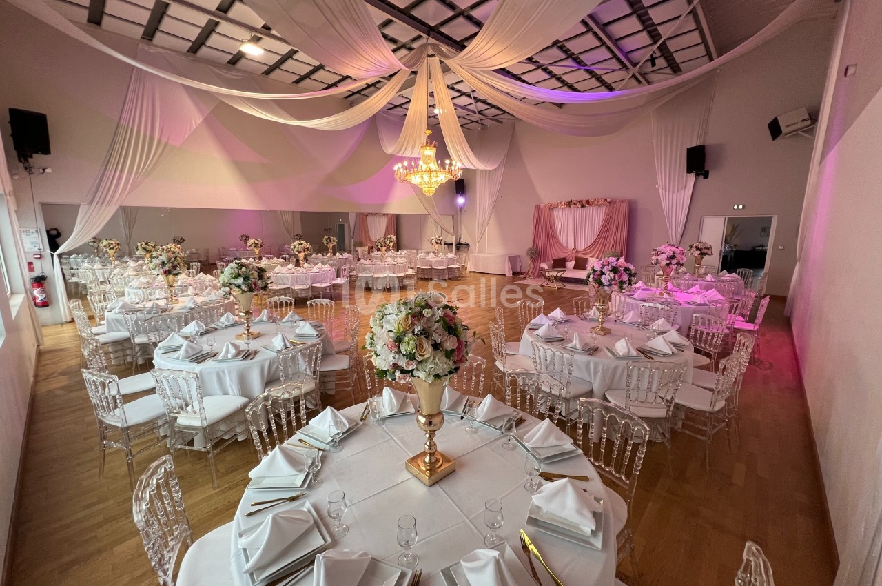 Salle de réception décorée pour un mariage, avec tables rondes dressées, chaises transparentes et plafonds ornés de drapés.