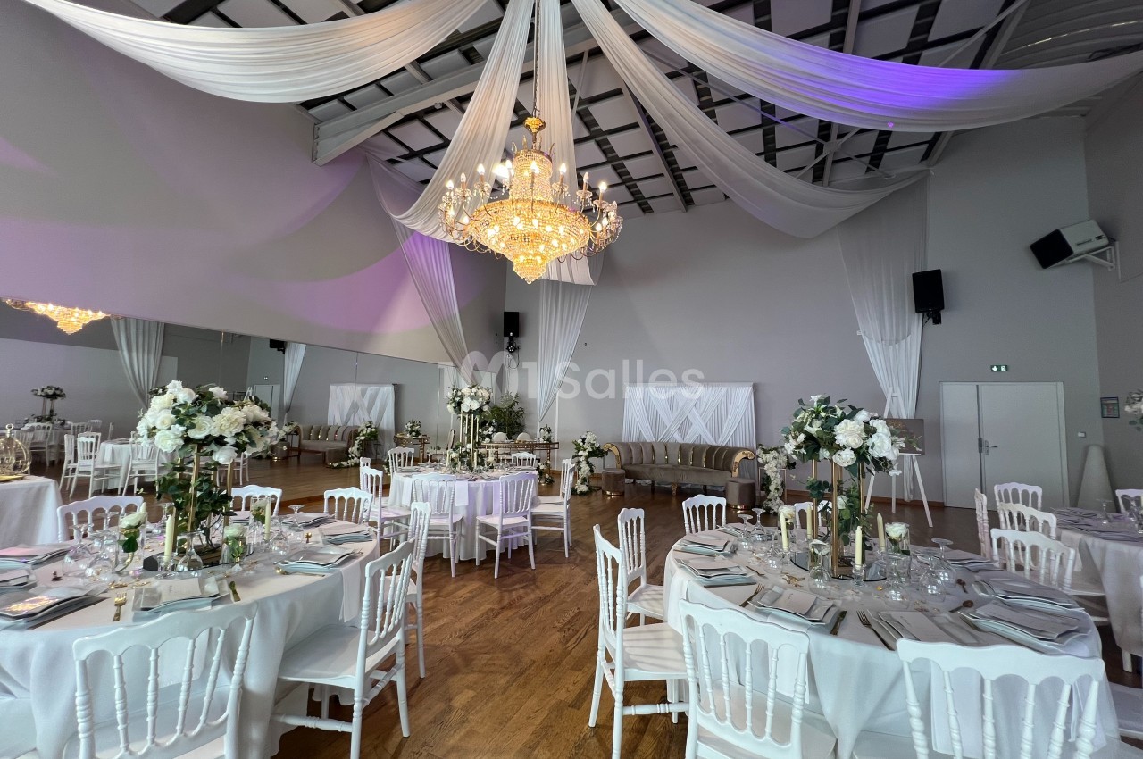 Salle de réception élégante avec tables dressées, chaises blanches, décor floral et grand lustre central.