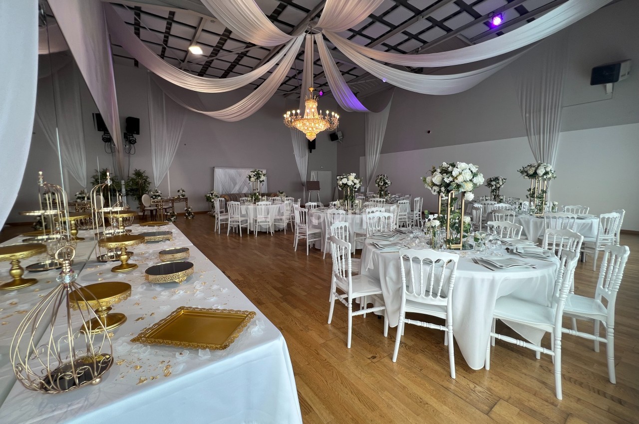 Salle de réception décorée avec des tables rondes, des chaises blanches, des fleurs et des drapés suspendus au plafond.