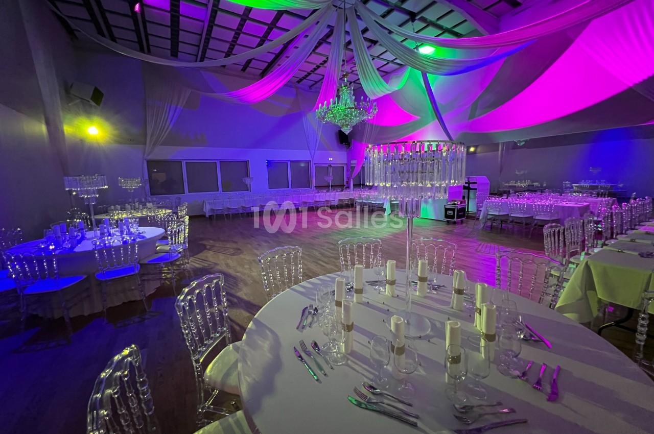 Salle de réception décorée avec des tables dressées, des chaises transparentes et un éclairage coloré.