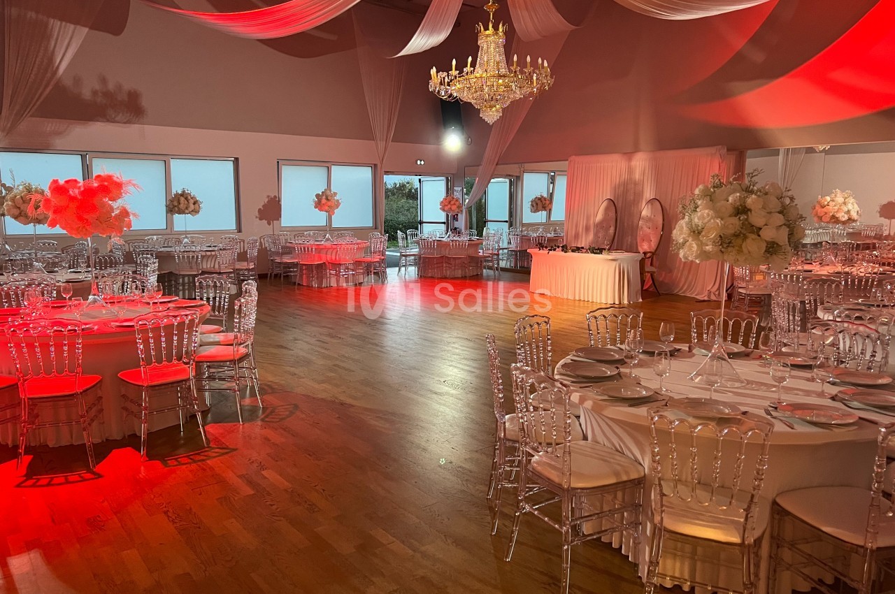 Salle de réception décorée avec des tables rondes, chaises transparentes, éclairage rouge et un grand lustre central.