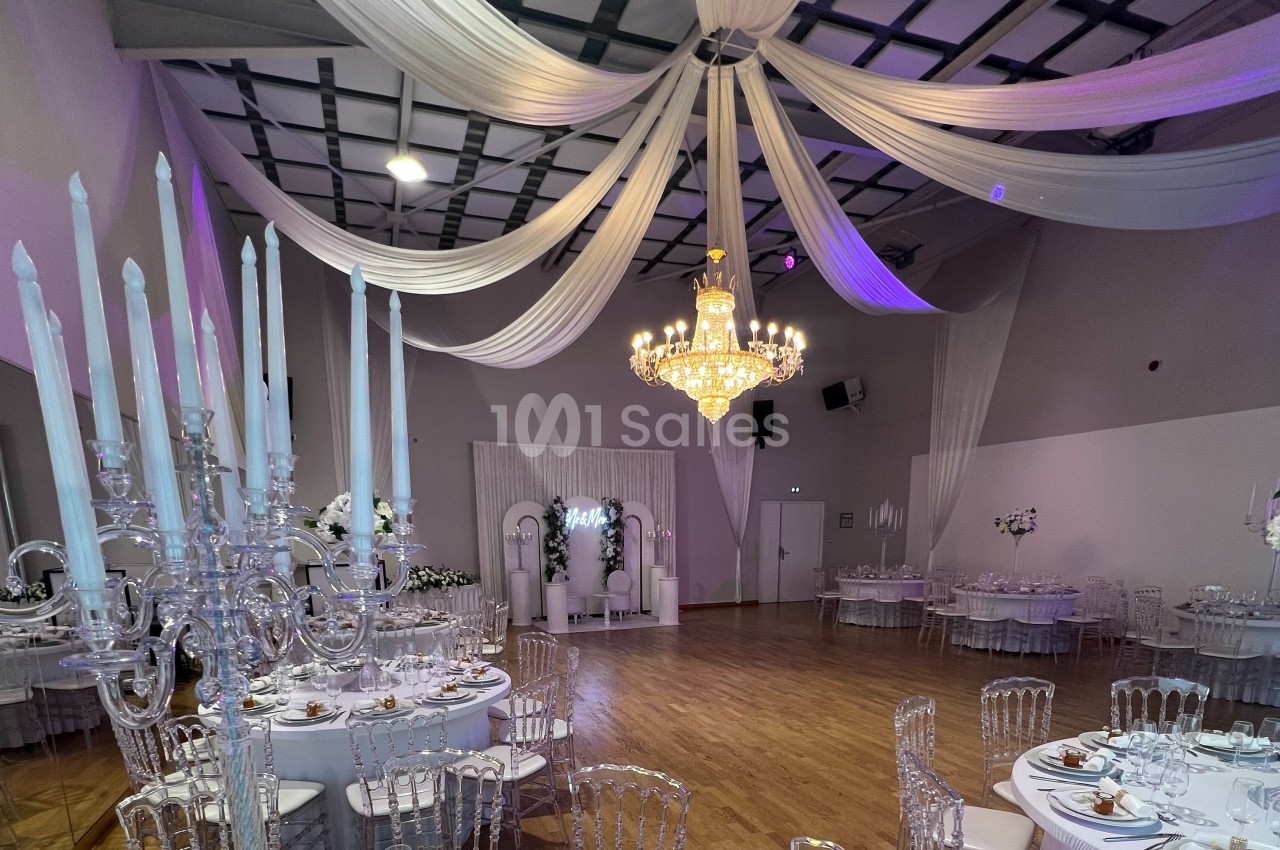 Salle de réception décorée avec des chandeliers, des tables rondes dressées et un grand lustre central.