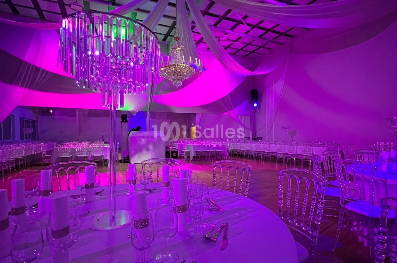 Salle de réception décorée avec des tables dressées, des chaises transparentes et un éclairage coloré violet et vert.