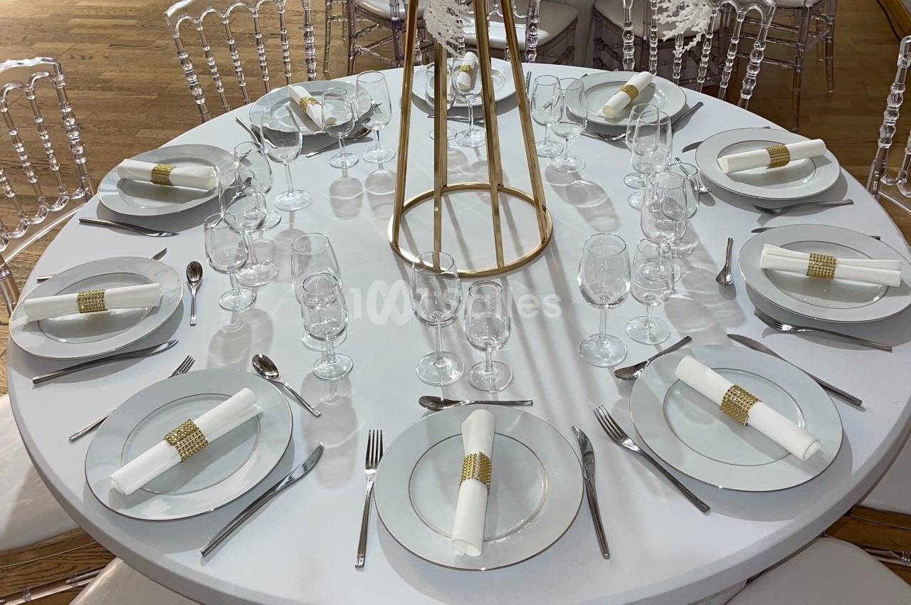 Table ronde élégamment dressée avec nappes blanches, assiettes, couverts, verres et serviettes ornées d'anneaux dorés.
