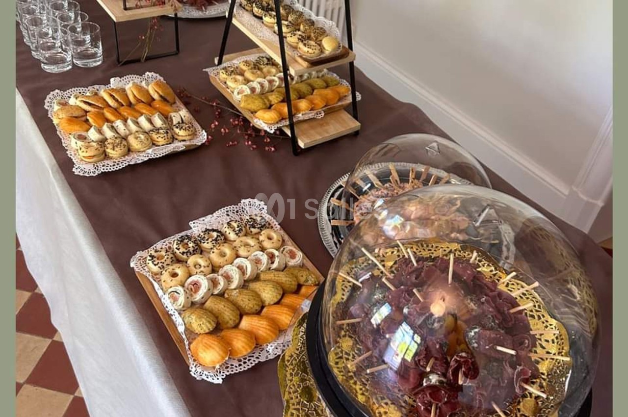 Table garnie de pâtisseries variées, verrines et charcuteries sous cloche, disposées sur une nappe marron.