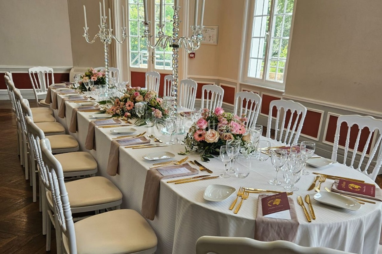Table élégamment dressée avec nappes blanches, chaises assorties, chandeliers, fleurs roses et couverts dorés.
