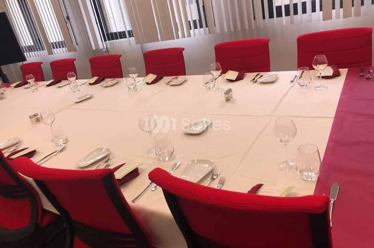 Table dressée avec nappes blanches, chaises rouges, verres, assiettes et serviettes, dans une salle lumineuse avec stores.