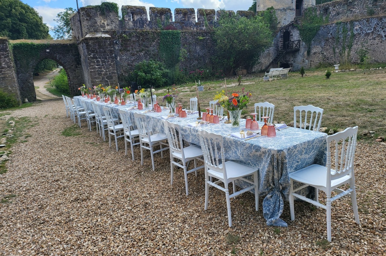 Grande table extérieure dressée avec nappes bleues, chaises blanches et décorations florales, devant des murs de pierre.