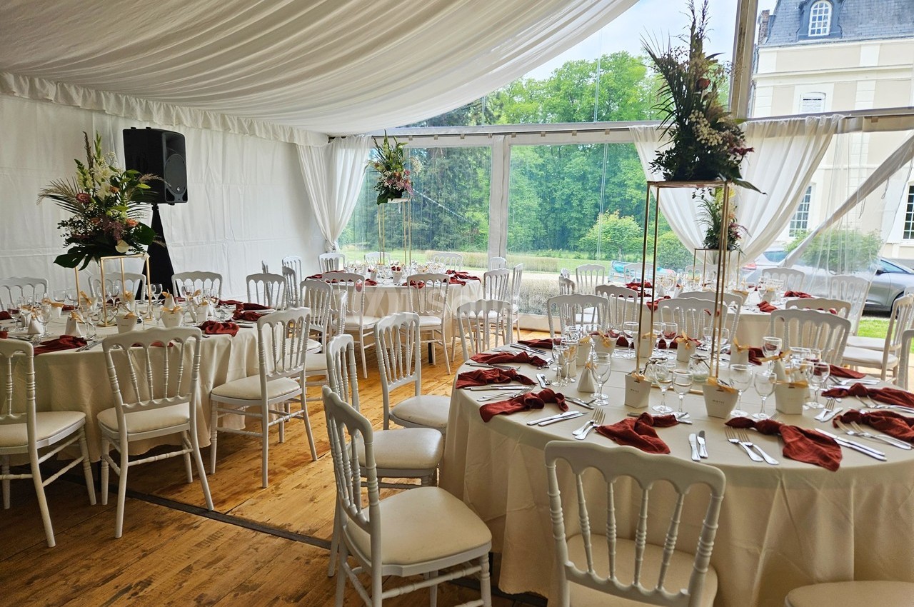 Salle de réception élégante avec tables rondes dressées, nappes blanches et serviettes rouges, vue sur un jardin verdoyant.