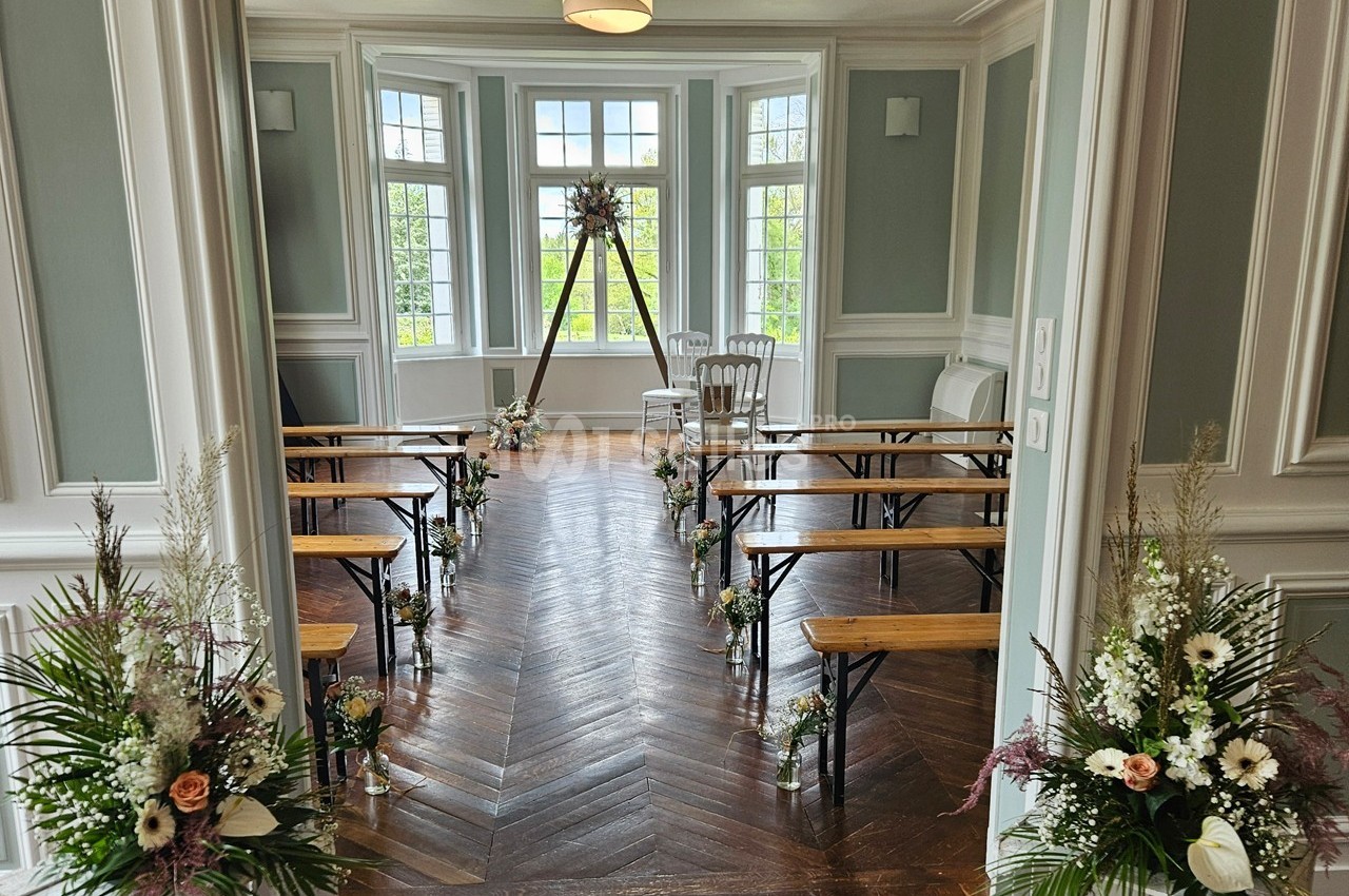 Salle lumineuse aménagée pour une cérémonie, avec bancs en bois, fleurs décoratives et grande fenêtre en arrière-plan.