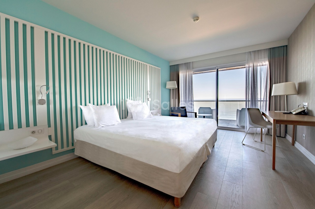 Chambre lumineuse avec lit double, bureau, fauteuils et grande baie vitrée offrant une vue sur la mer.